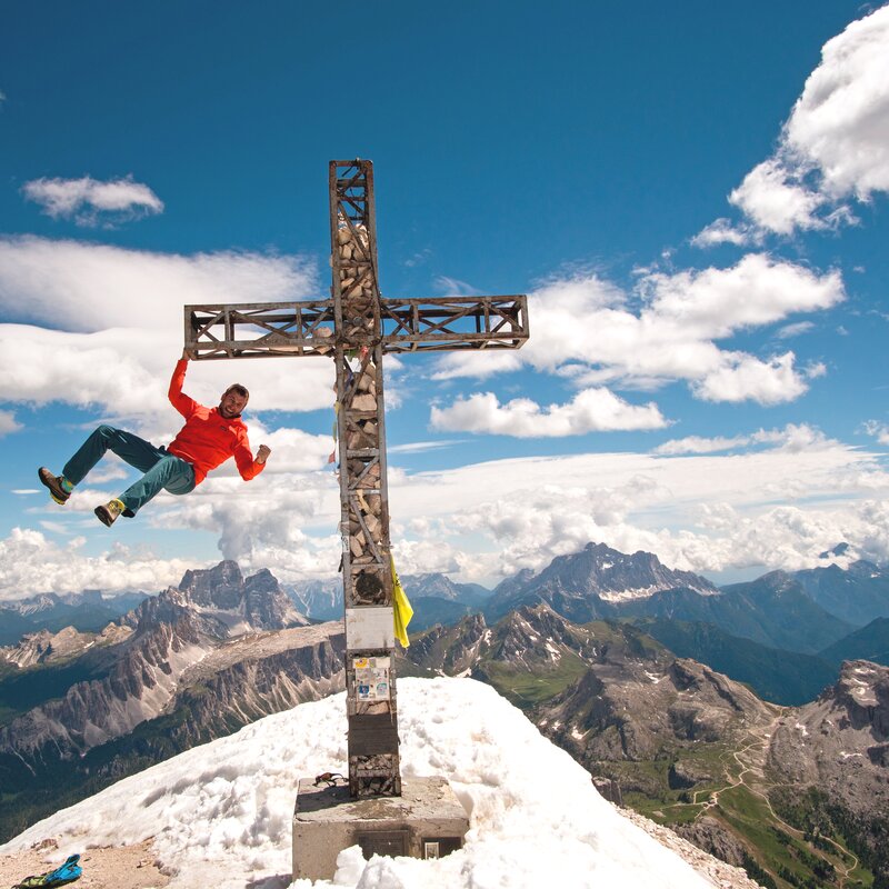 Arrampicatore appeso a una croce di vetta su una cima innevata con ampio panorama. | © Baumgartner Martin