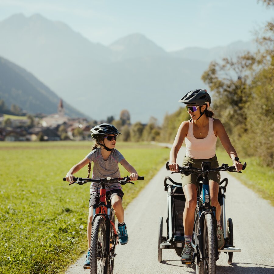 Madre e figlio pedalano fianco a fianco con un rimorchio per bambini e Rasun di Sopra sullo sfondo. | © IDM Südtirol - Alex Molling