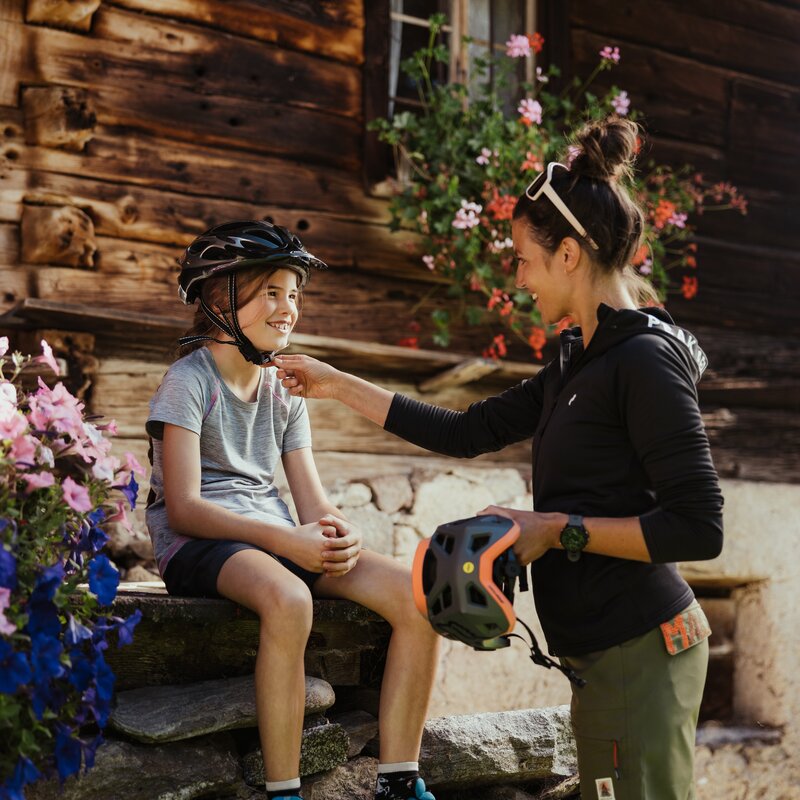 Una donna aiuta una bambina a mettere il casco da bici davanti a un maso antico con fiori. | © IDM Südtirol - Alex Molling