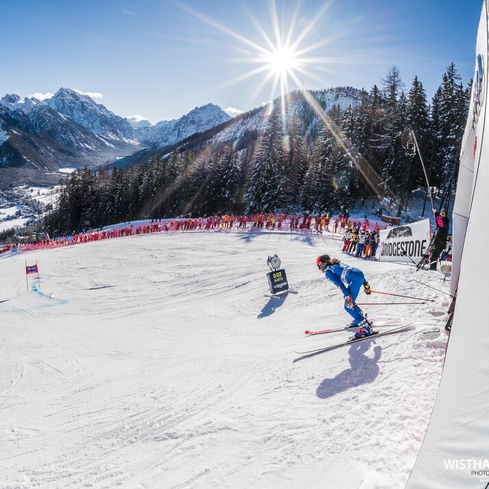 Eine Skirennläuferin startet den Riesenslalom und wird von Zuschauern am Rande der Piste bejubelt, im Hintergrund das Bergpanorama und strahlend blauer Himmel. | © Harald Wisthaler