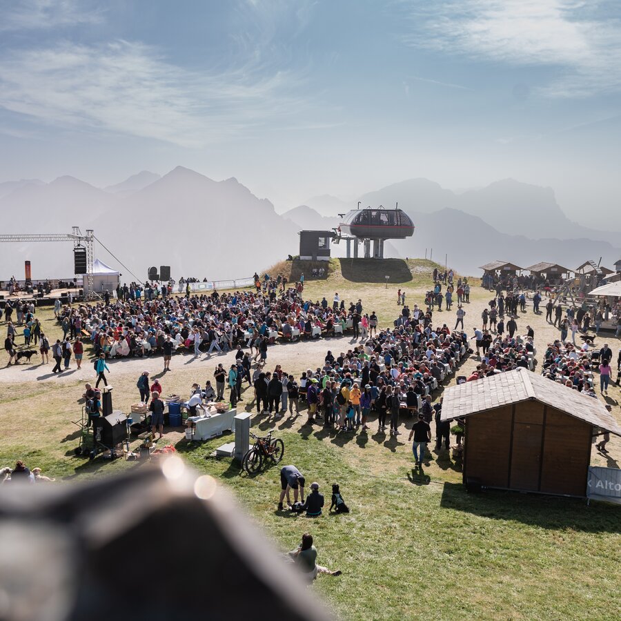 Zahlreiche Menschen versammeln sich auf einer Wiese vor Bühne und Marktständen mit Bergkulisse im Hintergrund. | © Gianvito Coco