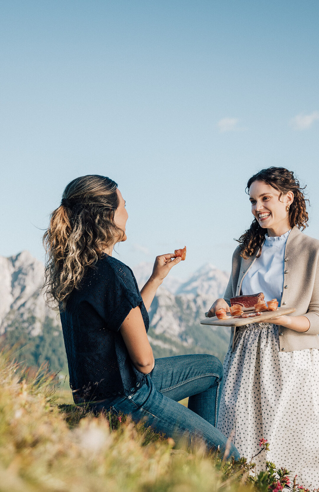 Vor dem Bergpanorama sieht man eine stehende Frau mit einem Brett Speck in den Händen und sie lächelt eine sitzende Frau an, welche sich an dem Speck bedient. | © IDM Südtirol