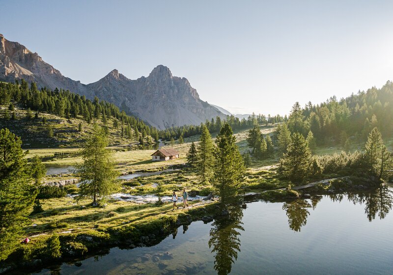 Der stille Bergsee spiegelt Nadelbäume und eine gezackte Bergkette bei klarem Morgenlicht. | © Harald Wisthaler