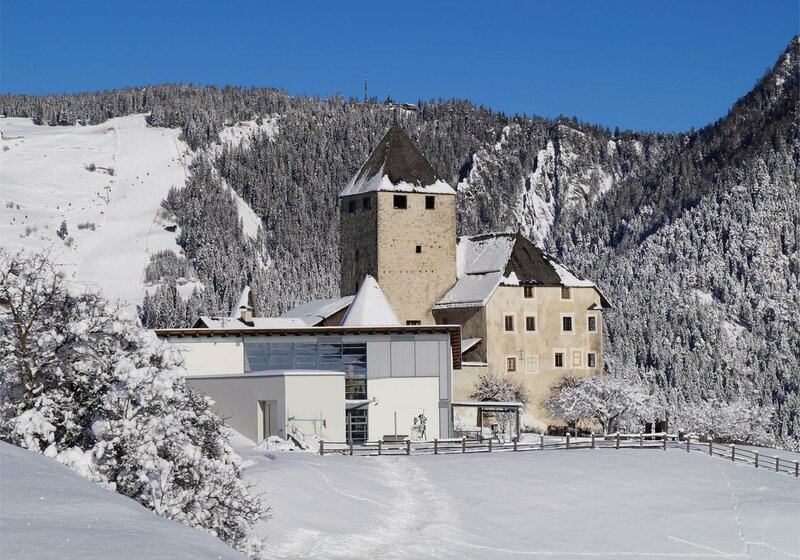 Eine verschneite Burganlage steht vor schneebedeckten Bergen unter blauem Himmel. | © Museum Ladin