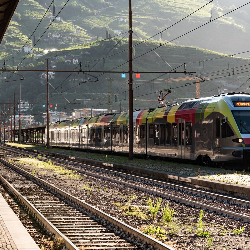 Zwei Züge stehen nebeneinander im Bahnhof mit Blick auf die umliegenden Berghänge. | © IDM Südtirol