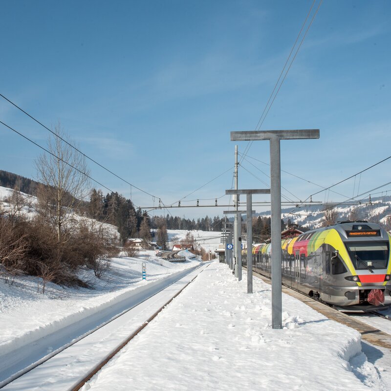 Zug hält bei Bahnhof in einem Dorf welches winterlich verschneit ist.