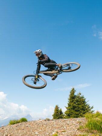 Eine Person auf einem Mountainbike fliegt durch die Luft, unter ihm ein Schotterweg und im Hintergrund Bäume und der blaue Himmel. | © Daniel Niederkofler