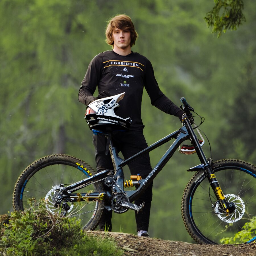 Ein junger Mann steht mit seinem Mountainbike im Wald, er hält seinen Helm in der Hand und blickt in die Kamera. | © Daniel Niederkofler