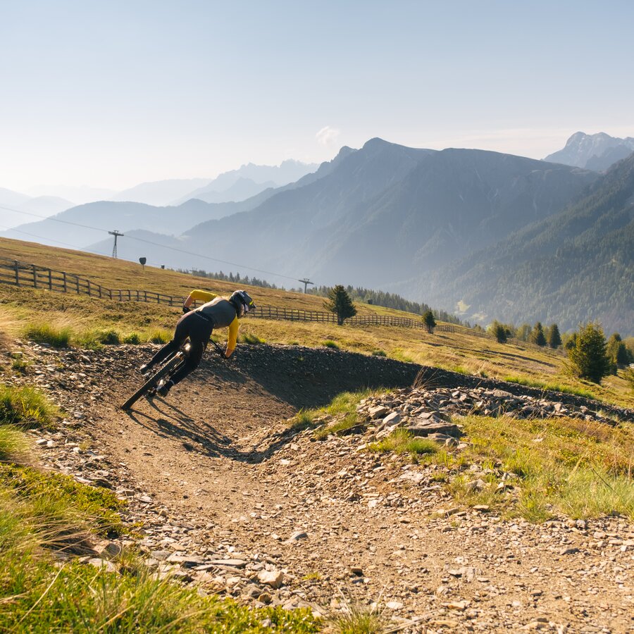 Eine Mountainbikerin fährt in die Kurve gelehnt einen schmalen Pfad am Berg entlang, im Hintergrund das Bergpanorama. | © Daniel Niederkofler