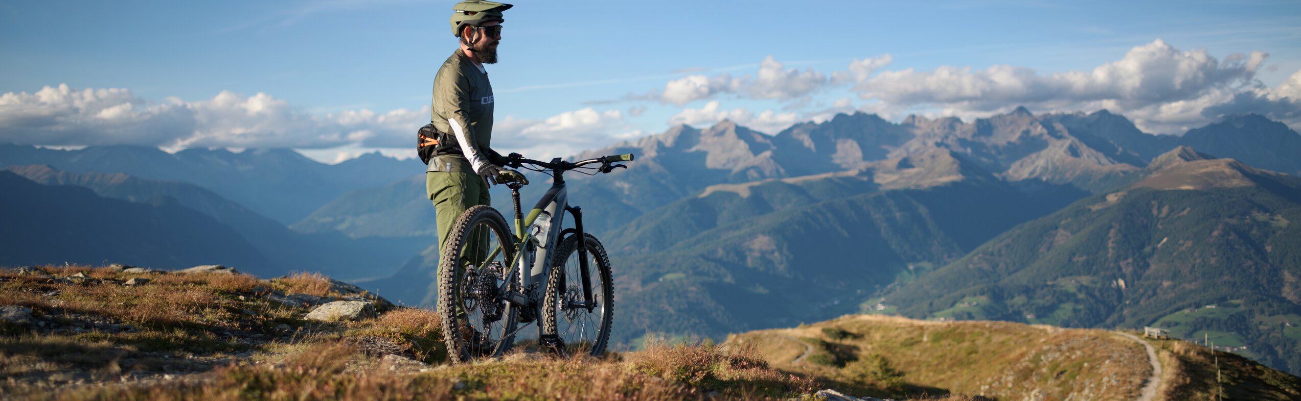 Ein Biker steht mit dem Fahrrad an einem Berghang und blickt über eine weite Gebirgslandschaft. | © Cube Bikes