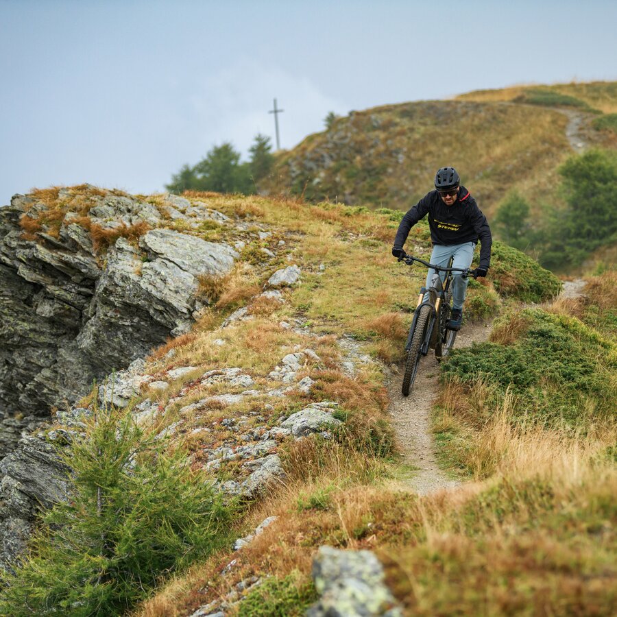Ein Mountainbiker fährt bergab auf einem schmalen Trail entlang eines Kamms, im Hintergrund ein Gipfelkreuz. | © Cube Bikes