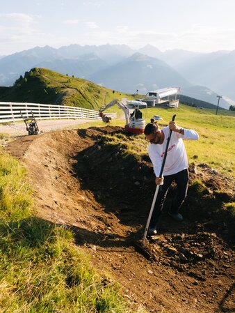 Eine Person bearbeitet mit einer Harke einen Erdhügel auf einem Mountainbike-Trail in Hanglage. | © Daniel Niederkofler