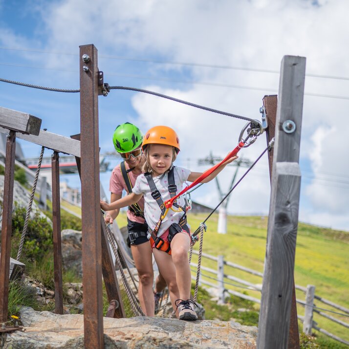 Ein kleines Mädchen in Kletterausrüstung klettert durch einen kleinen Parcours, dicht hinter ihr eine lächelnde Frau und im Hintergrund eine Bergwiese und der leicht bewölkte Himmel. | © Gianvito Coco