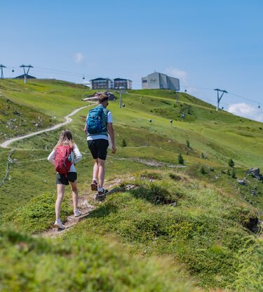 Zwei Personen wandern einen Wanderpfad entlang auf einen Berg, im Hintergrund Gebäude und Gondelbahnen. | © Gianvito Coco
