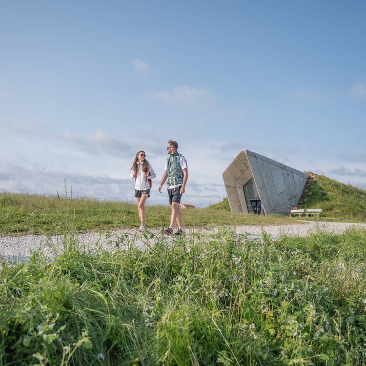 Zwei Wandernde laufen auf einem Wiesenpfad am modernen Aussichtspavillon vorbei. | © Gianvito Coco