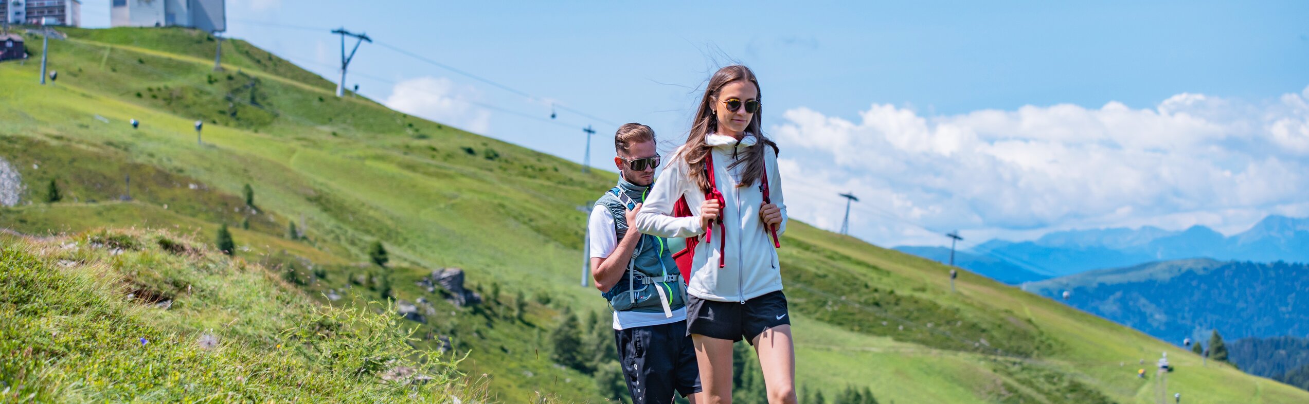 Ein Paar wandert über einen grünen Hang mit der Kronplatz-Infrastruktur im Hintergrund. | © Gianvito Coco