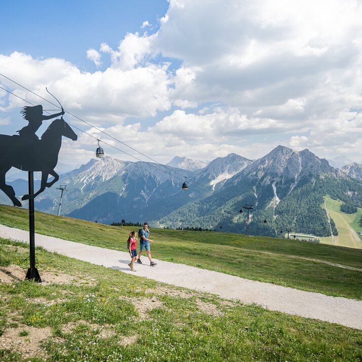 Ein Mann und eine Frau wandern im Sommer am Kronplatz vorbei an der Dolasilla Statue, die Dolomiten im Hintergrund. | © Harald Wisthaler