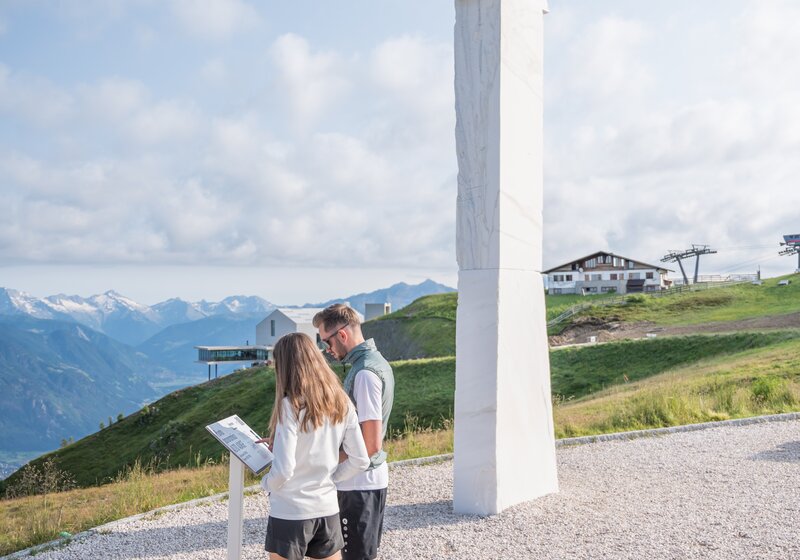 Zwei Personen stehen auf einem Berg neben einer Kunstskulptur und lesen sich das Beschreibungsschild durch, im Hintergrund der leicht bewölkte Himmel. | © Gianvito Coco
