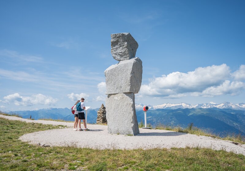 Zwei Personen stehen auf einem Berg vor einer großen Steinskulptur und lesen das Beschreibungsschild, im Hintergrund weitere Berge und der leicht bewölkte Himmel. | © Gianvito Coco