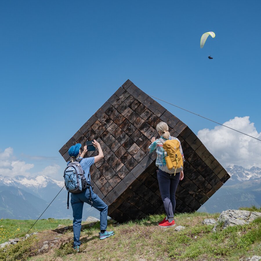 Zwei Personen stehen vor einem großen würfelförmigen Holzkunstwerk auf dem Berg, im Hintergrund das Bergpanorama, leicht bewölkter Himmel und ein Paraglider. | © Gianvito Coco