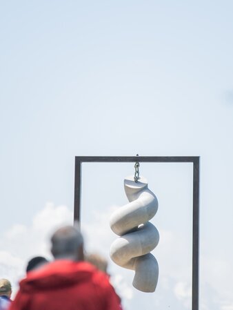 Ein in einer Metallhalterung hängendes Betonkunstwerk hängt vor dem Hintergrund des strahlend blauen Himmels und einigen Menschen.  | © Gianvito Coco