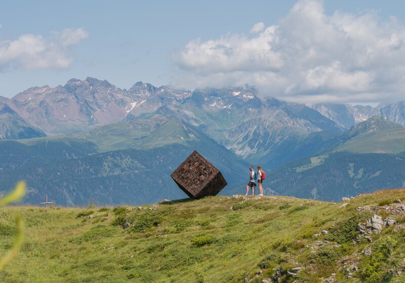 Zwei Personen stehen neben einer riesigen hölzernen würfelförmigen Kunstskulptur auf einem Berg, im Hintergrund weitere Berge. | © Gianvito Coco