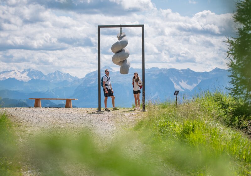 Zwei Personen stehen unter einem hängenden Kunstwerk auf einem Berg, im Hintergrund das Bergpanorama und der leicht bewölkte Himmel. | © Gianvito Coco