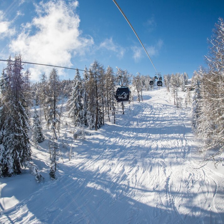 Eine Gondelbahn fährt durch einen Wald, darüber Sonne, einzelne Wolken und ansonsten blauer Himmel. | © Harald Wisthaler