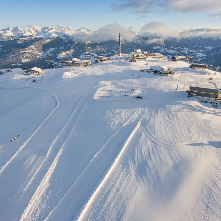 Luftaufnahme eines sonnigen Gipfelplateaus mit präparierten Skipisten, Gebäuden und umliegender Berglandschaft. | © Harald Wisthaler