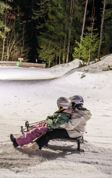 Zwei Menschen rodeln lachend eine schneebedeckte, kurvige Waldpiste hinab. | © Gianvito Coco