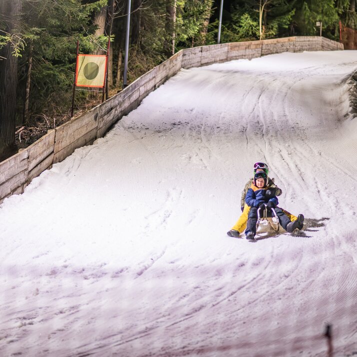 Ein Kind fährt auf einem Schlitten eine beleuchtete Nachtrodelbahn im Wald hinunter. | © Gianvito Coco
