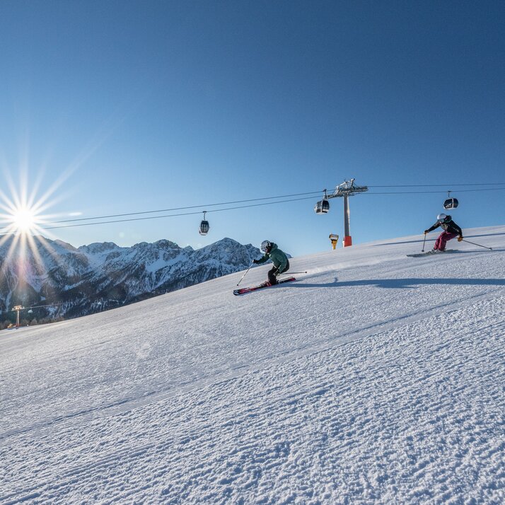 Zwei Skifahrer auf einer Piste, im Hintergrund eine Gondel, Berge, Sonne und blauer Himmel. | © Skirama Kronplatz